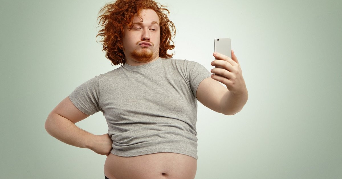 Funny overweight plump man with duck lips wearing undersize t-shirt with belly hanging out of pants, keeping hand on waist, posing for selfie, holding cell phone, trying to seem attractive and sexy